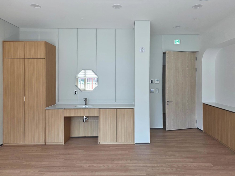 Patient room with pale wall panels, an oak wardrobe and vanity unit with a sink, an octagonal mirror and an oak door