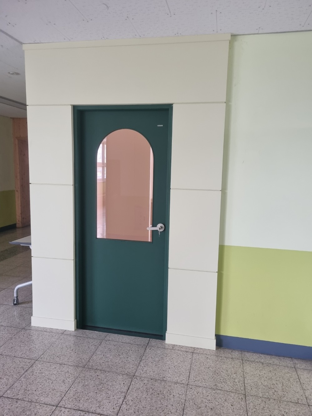 Dark green classroom door with an arched vision panel in a cream film surround, green wainscot band on the wall beside it