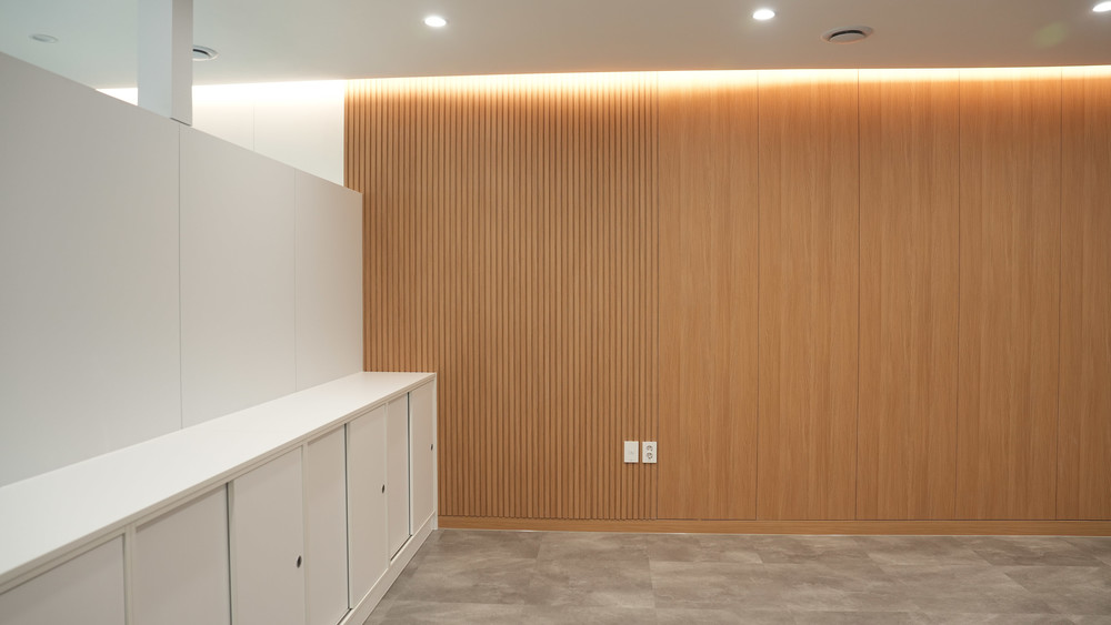 Oak slatted feature wall with cove lighting above it, white low cabinets in front, grey stone-look floor